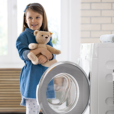 Young girl standing in front of an open tumble dryer, cuddling a warm teddy fresh from the machine. Links to Laundry Category Page.