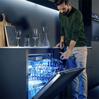 Man emptying a dishwasher with a blue interior light in a sleek, modern black kitchen. The purpose of the image id to link to the Dishwasher Category Page.