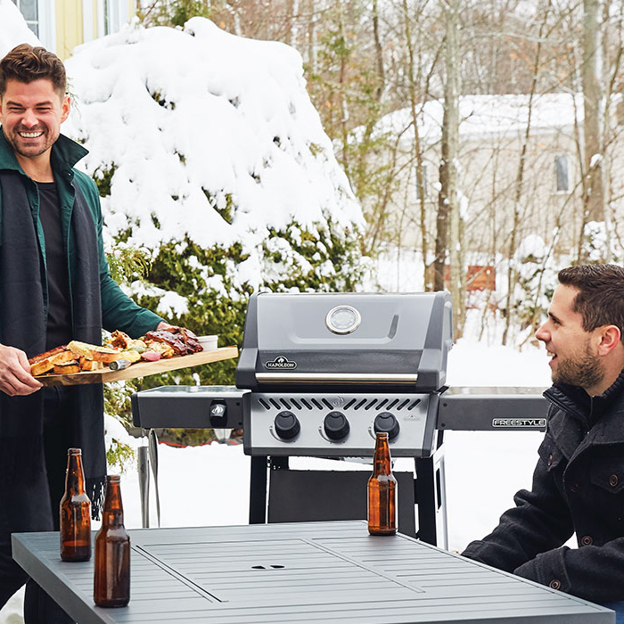 Group of friends enjoying a beer while preparing a Napoleon BBQ in snowy weather. Link to Home & Outdoors Category Page.